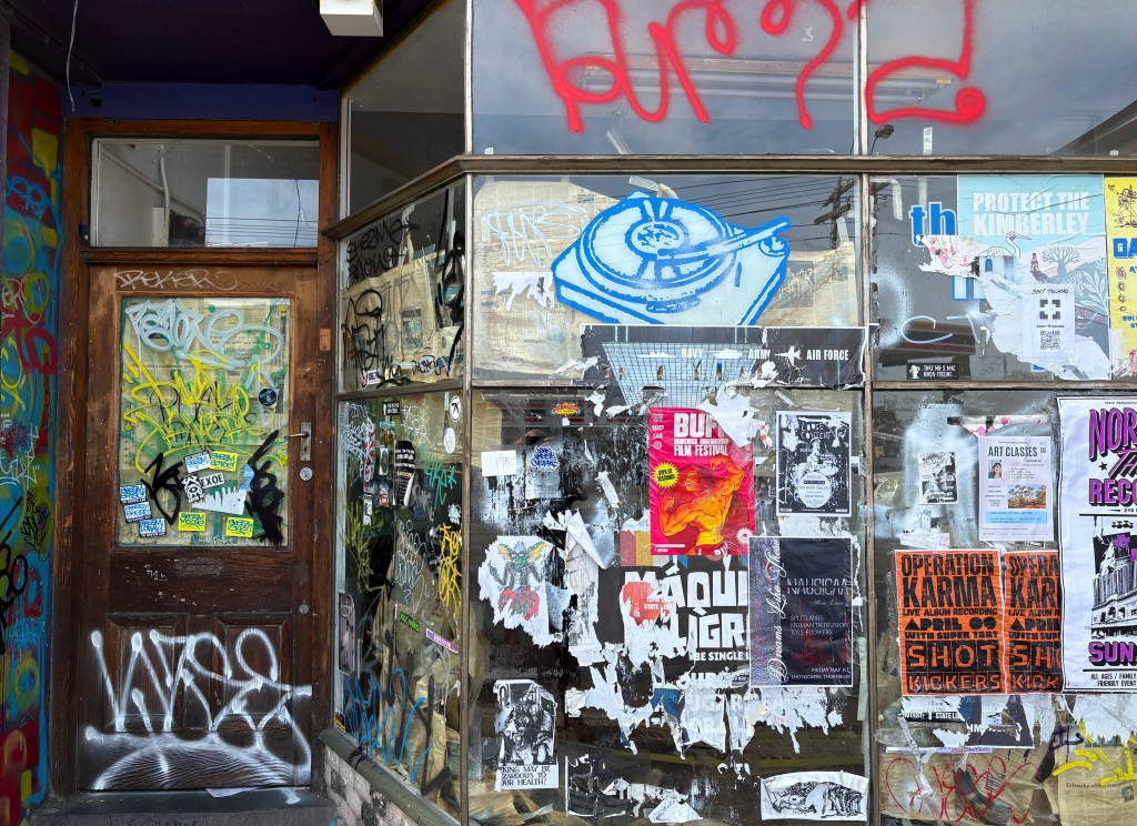 A graffiti-covered shopfront with a wooden door, featuring a mix of vibrant stickers and posters on the glass, showcasing various events and messages.