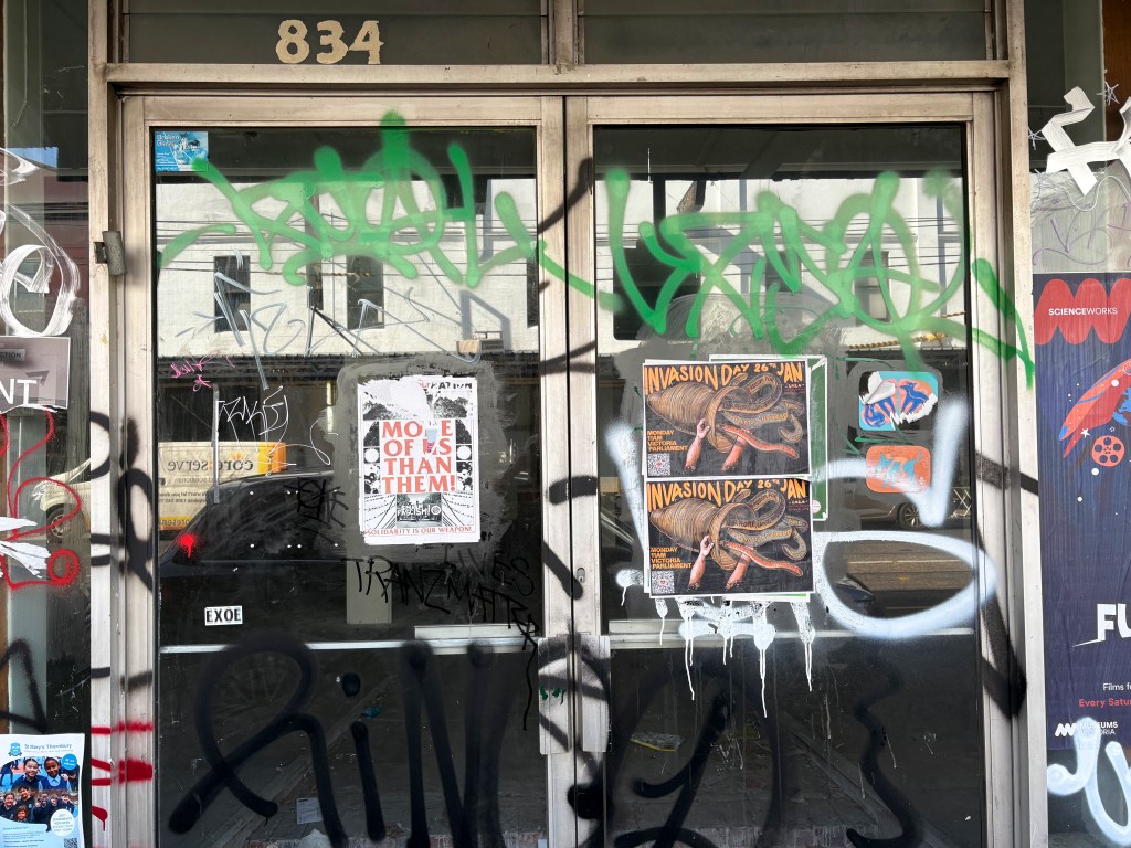 A storefront with the number 834, covered in various graffiti and street art. Posters for an event titled 'Invasion Day' and a film screening are partially visible behind the graffiti.