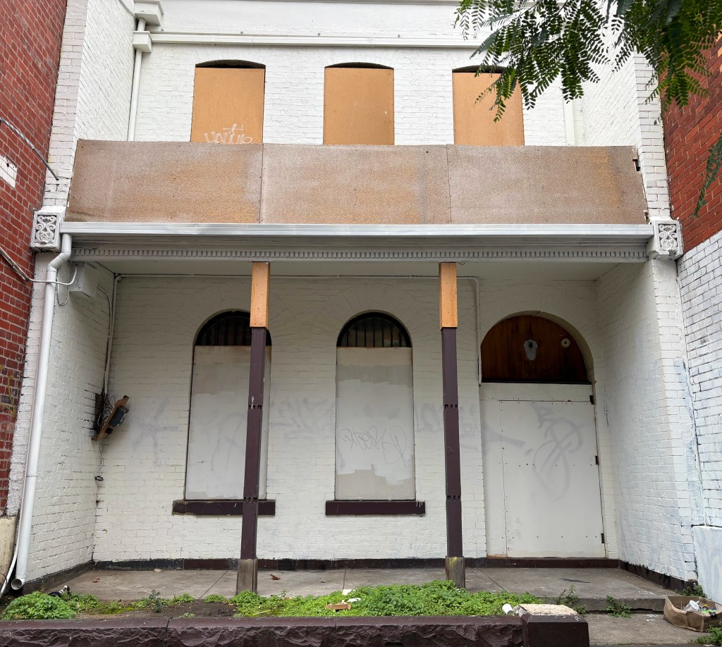 View of an abandoned building with boarded windows and a partially covered entrance, featuring white and red brick walls.