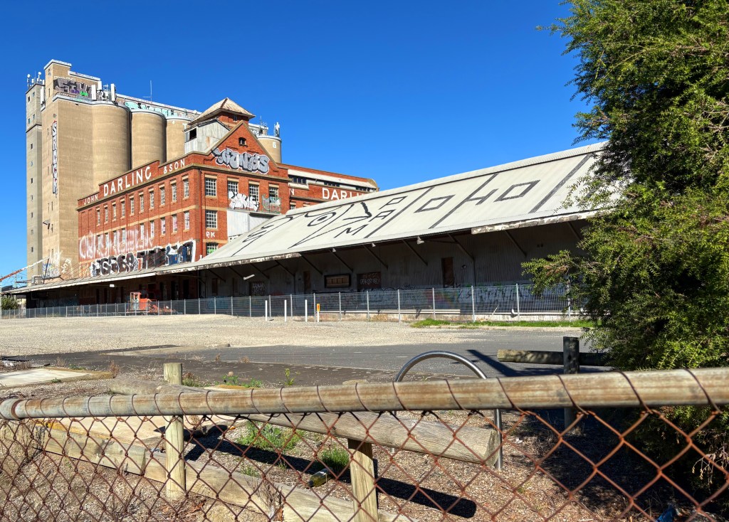 Abandoned industrial building with graffiti, featuring a storage facility and a large grain silo in the background, surrounded by fencing and vegetation under a clear blue sky.