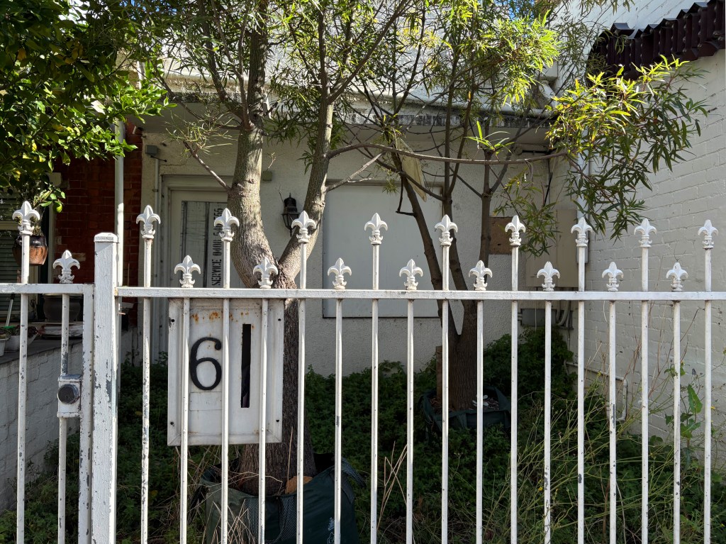 A white wrought iron fence surrounding a house with a decorative gate, featuring the number 61 prominently displayed. Two trees are visible behind the fence, alongside some overgrown plants.