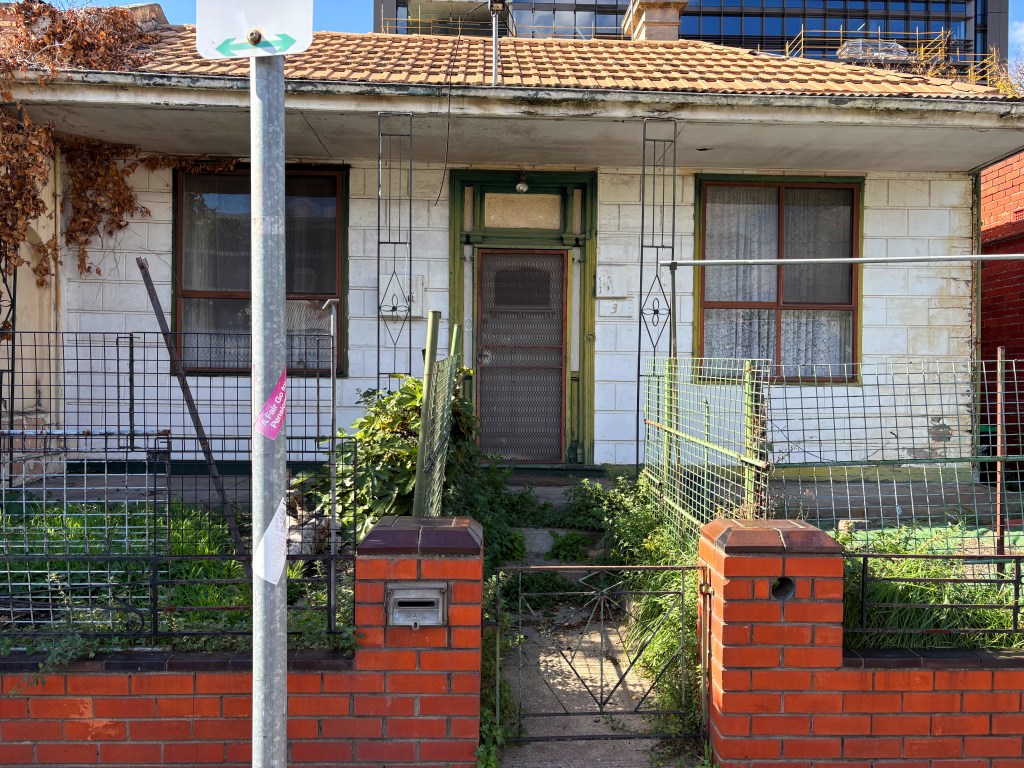 Front view of an old house with a porch and a green door, surrounded by overgrown plants and a wire fence. A street sign is visible in front of the property.