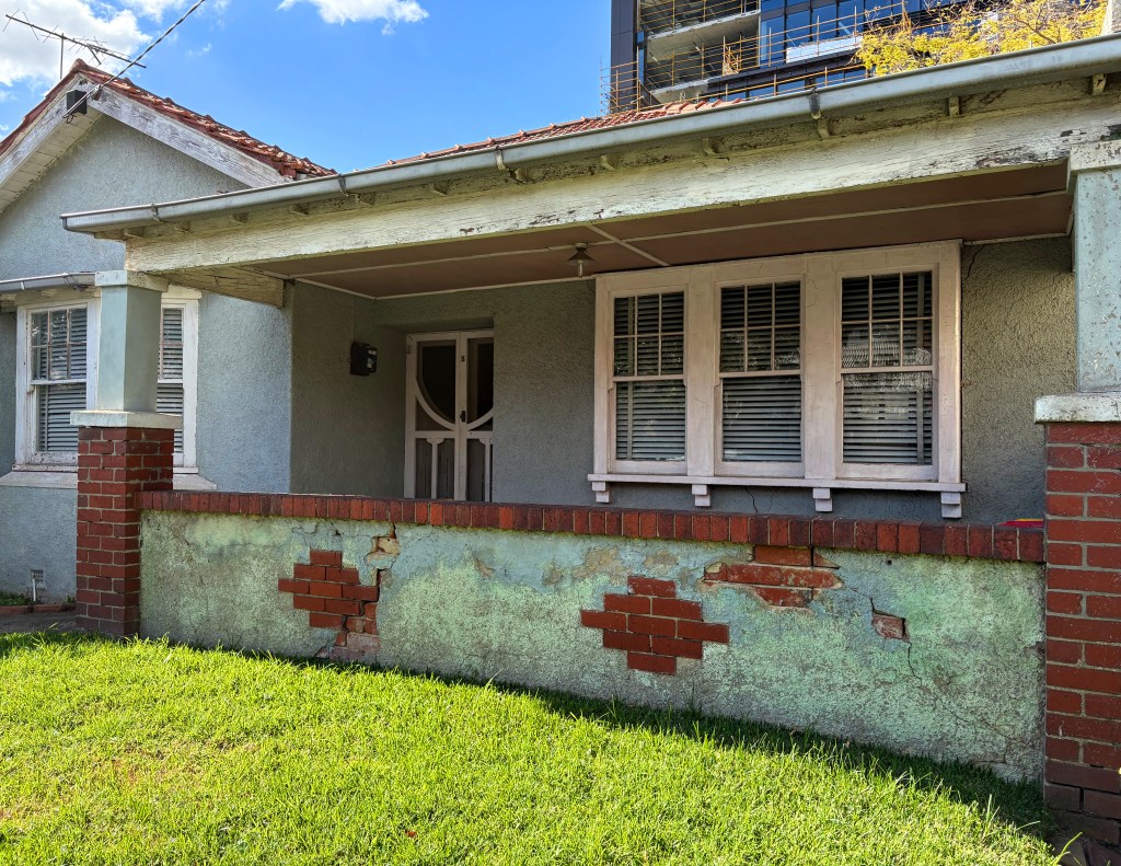 Front view of an old house featuring a porch with a sloped roof, classic brick columns, and a wall displaying peeling paint and exposed bricks, situated on a grassy lawn.
