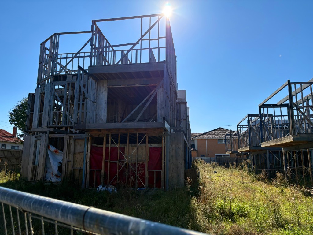 Construction site featuring a partially built wooden structure with framing exposed, surrounded by tall grass and adjacent buildings under construction, under a clear blue sky.