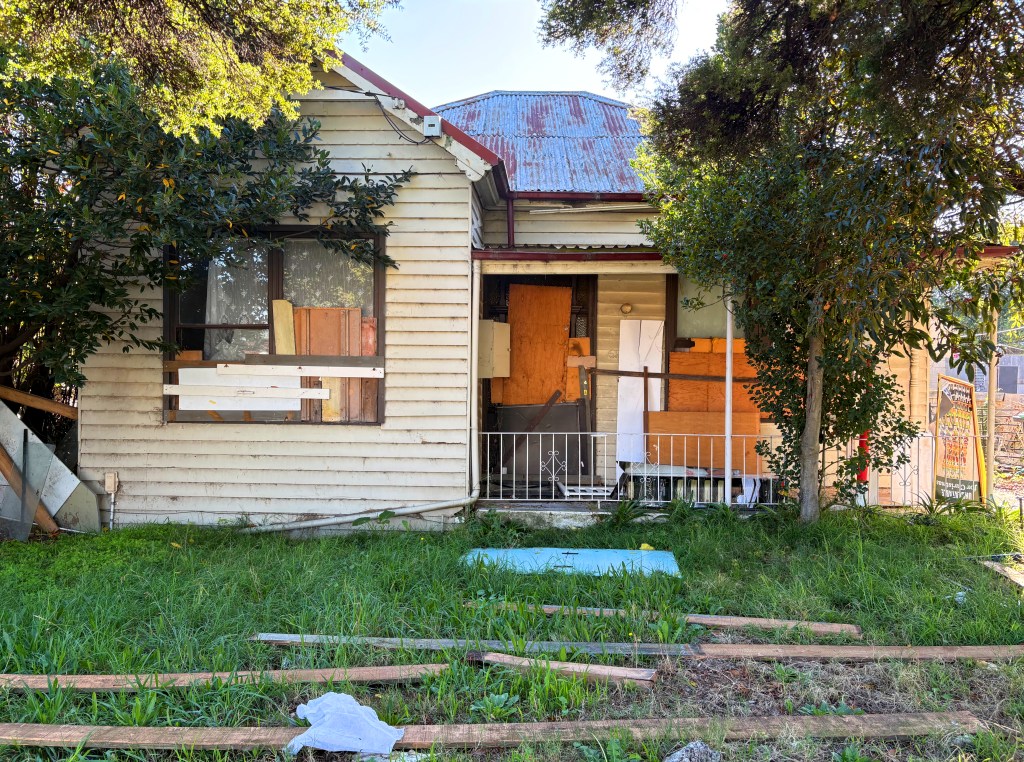 Front view of a dilapidated house with boarded windows, overgrown grass, and scattered wooden planks.