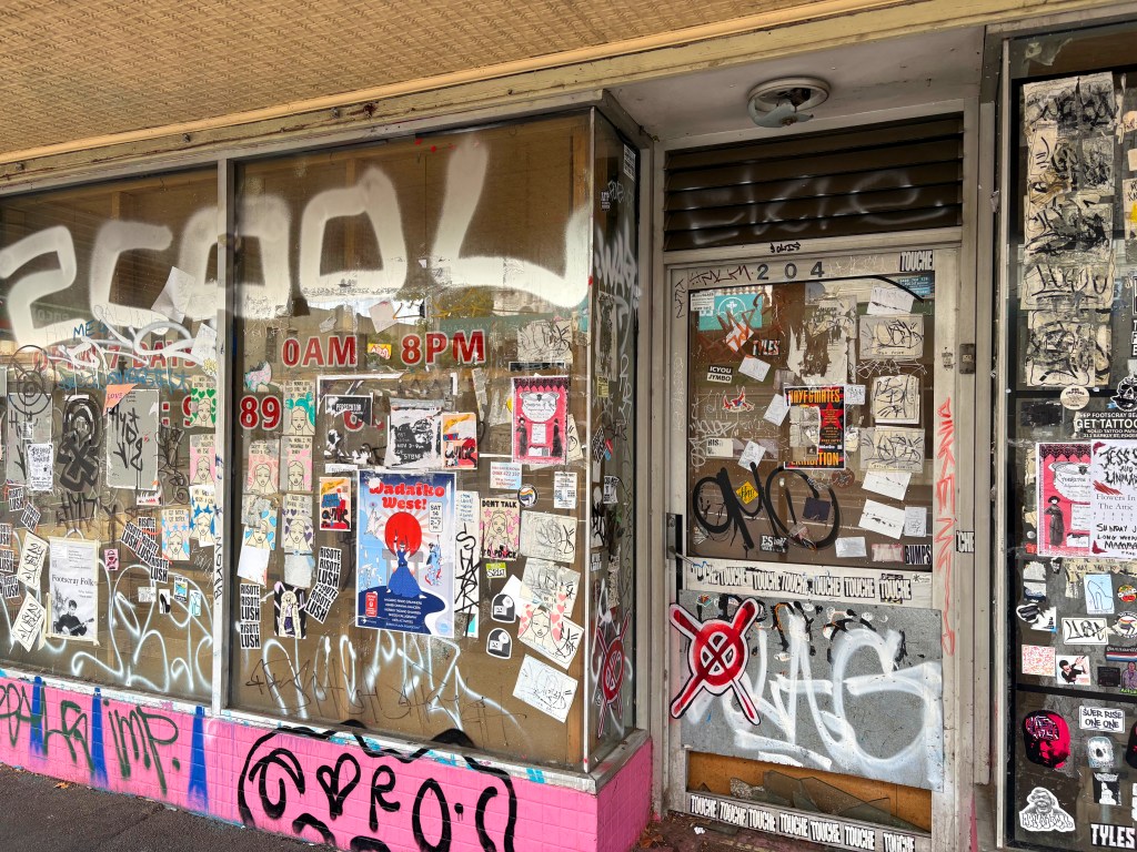 A graffiti-covered storefront with a glass window showcasing numerous posters and stickers, along with the words '0AM 8PM' and '89' prominently displayed.