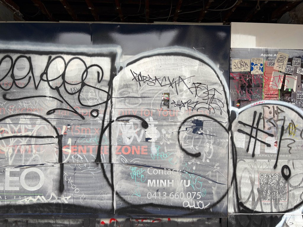 A wall covered in various types of graffiti, including tags, scribbles, and stickers, with some text partially visible underneath.