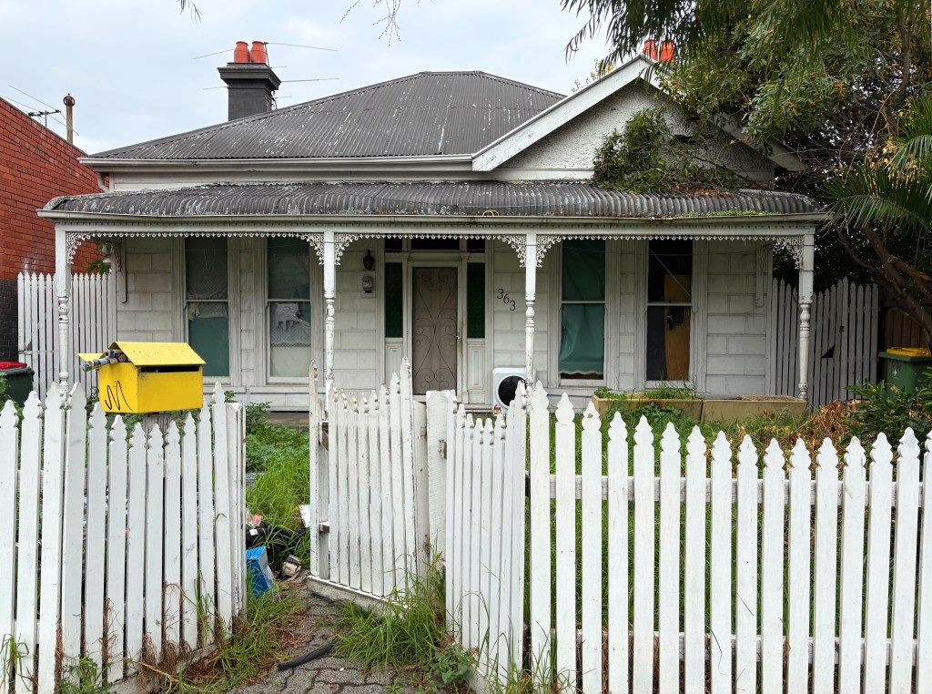 Front view of an abandoned, old house with a white picket fence. The house features a sloping roof, a small porch, and overgrown grass in the garden. A yellow mailbox is visible beside the entrance.