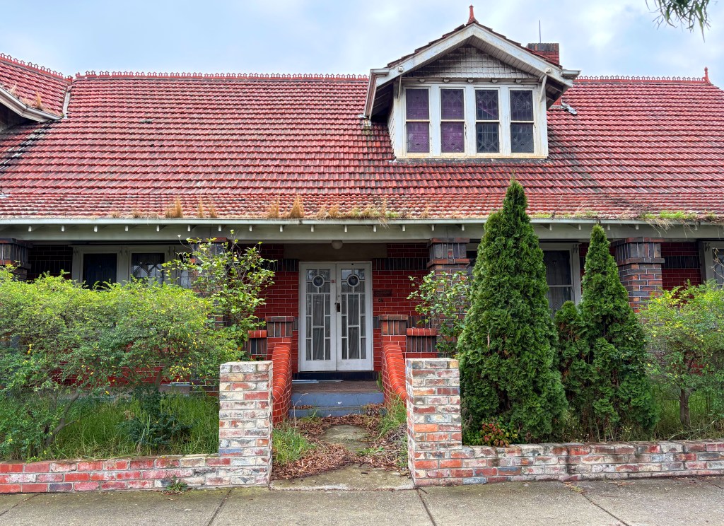 A front view of an old brick house with a red tiled roof, featuring two tall conifer trees on either side of the entrance, which has decorative double doors. Overgrown bushes and grass are visible in the garden, indicating neglect.