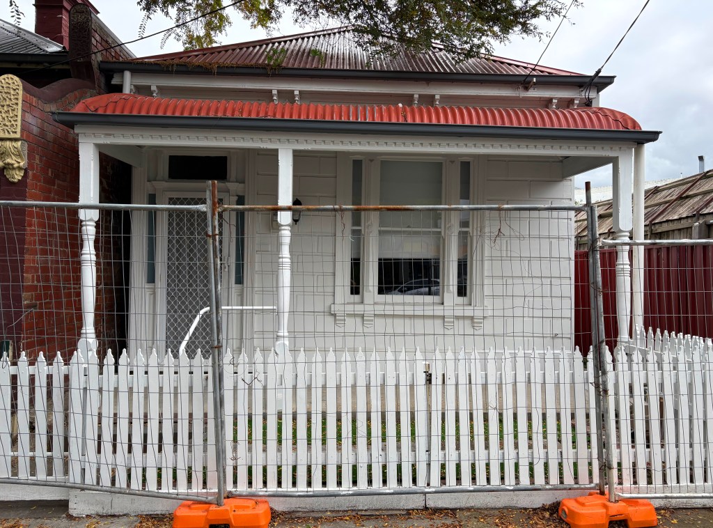 A white house with a red roof, partially enclosed by a construction fence and an orange safety barrier in the foreground.