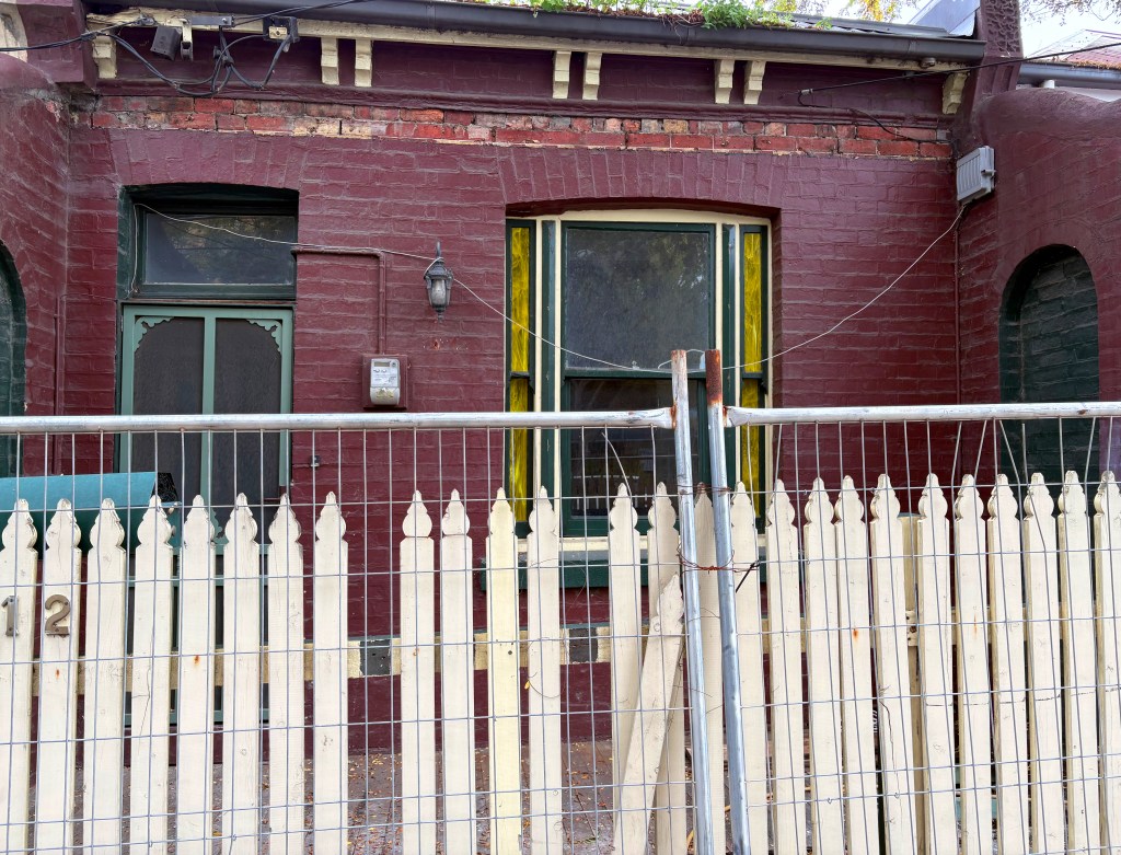 Front view of a red-brick house with a green door and a white picket fence, partially fenced off. The window has yellow accents and a lantern beside the entrance.