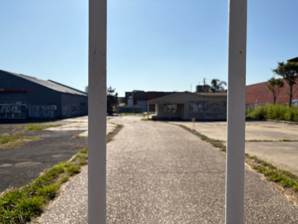 View of an abandoned lot through a fence, with buildings in the background and a clear blue sky.