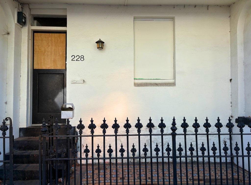 Front entrance of a building featuring a wooden door with a large piece of plywood covering a window. The house number 228 is displayed beside the door, and a small mailbox is mounted nearby. A decorative black iron fence encloses the steps leading up to the door.