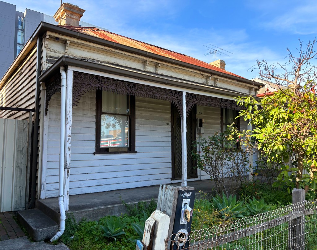 A weathered single-storey wooden house with a red corrugated iron roof, decorative verandah elements, and a small garden in front, surrounded by a fence.