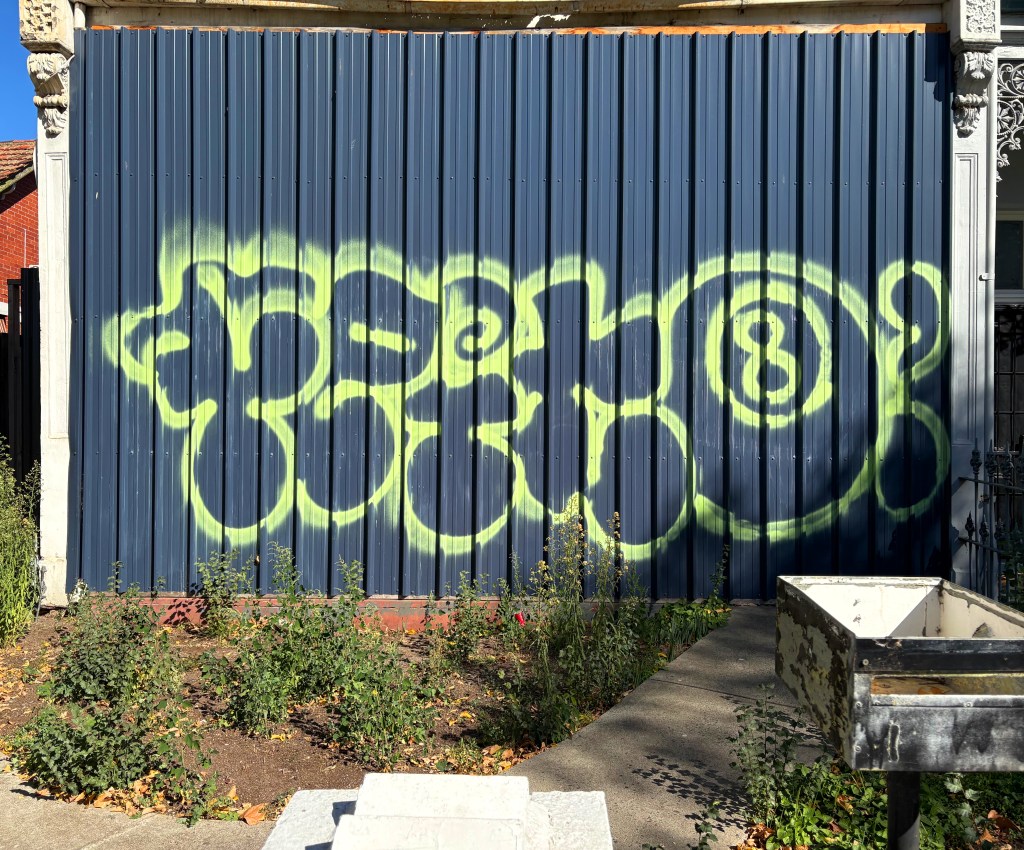 A vibrant green graffiti artwork on a blue corrugated metal wall, featuring abstract shapes and swirls, set in a garden area.