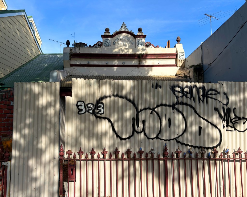 A heritage building with decorative features, partially obscured by a metal fence covered in graffiti. The blue sky is visible above.