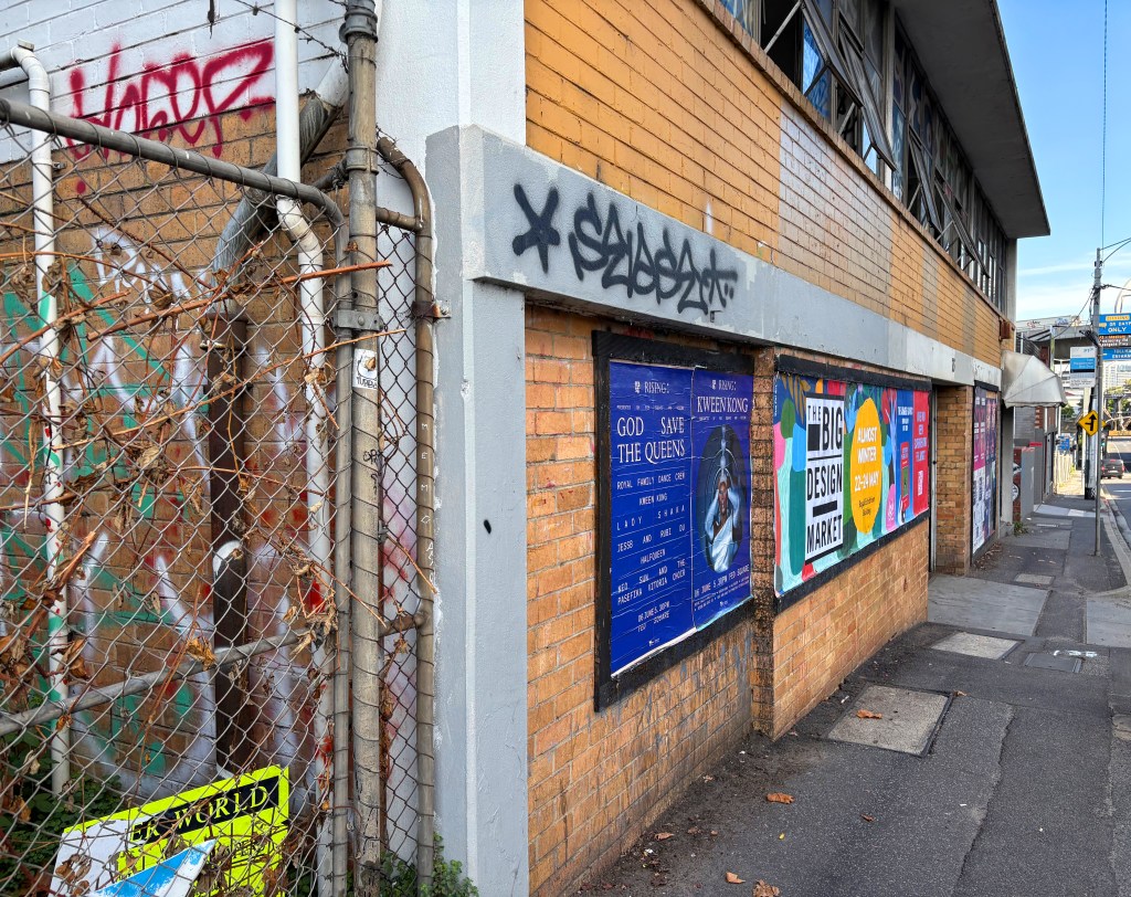 A street view of a brick building with graffiti on its walls and a chain-link fence. Posters are displayed on the building, advertising events, while the sidewalk shows some discarded signage and overgrown plants.