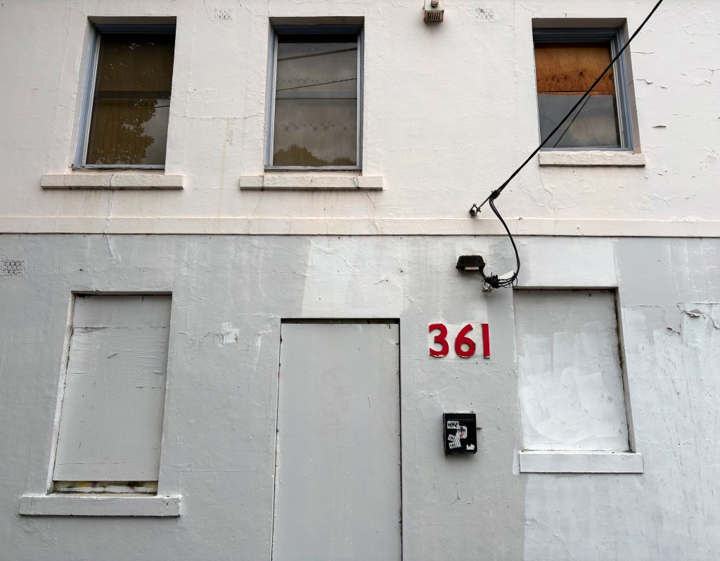 Exterior view of a weathered building with two windows, one boarded up, and a door underneath a large red house number '361'.