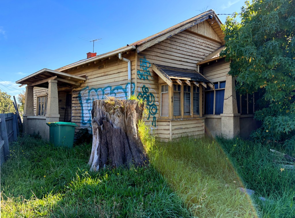 Abandoned house with wooden exterior and graffiti, surrounded by overgrown grass and a tree stump.