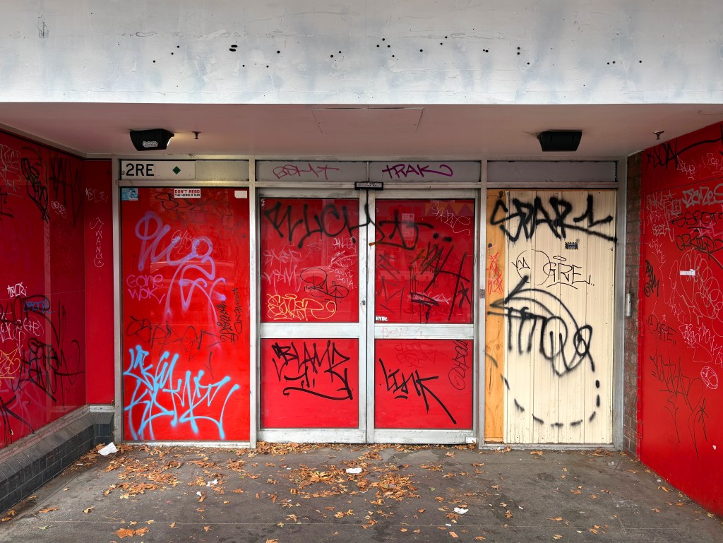 A building facade with three doors, covered in colourful graffiti and tags. The walls are primarily red, with scattered leaves on the ground.