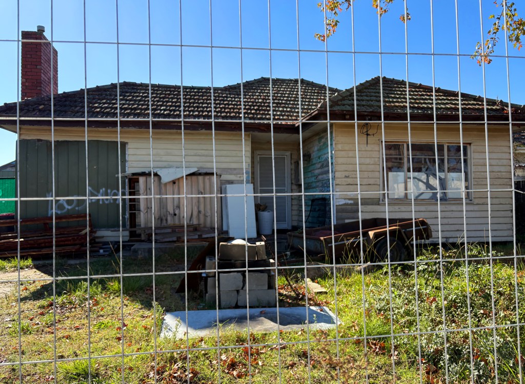 An old, weathered house behind a wire fence, featuring a chimney, uneven siding, and graffiti. The yard is overgrown with grass and debris scattered around.