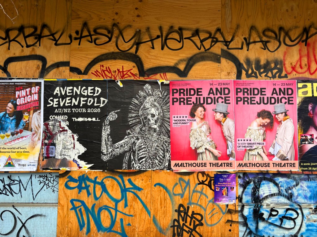 A wall covered in graffiti with various posters including one for Avenged Sevenfold's tour, and two for a performance of Pride and Prejudice at the Malthouse Theatre.
