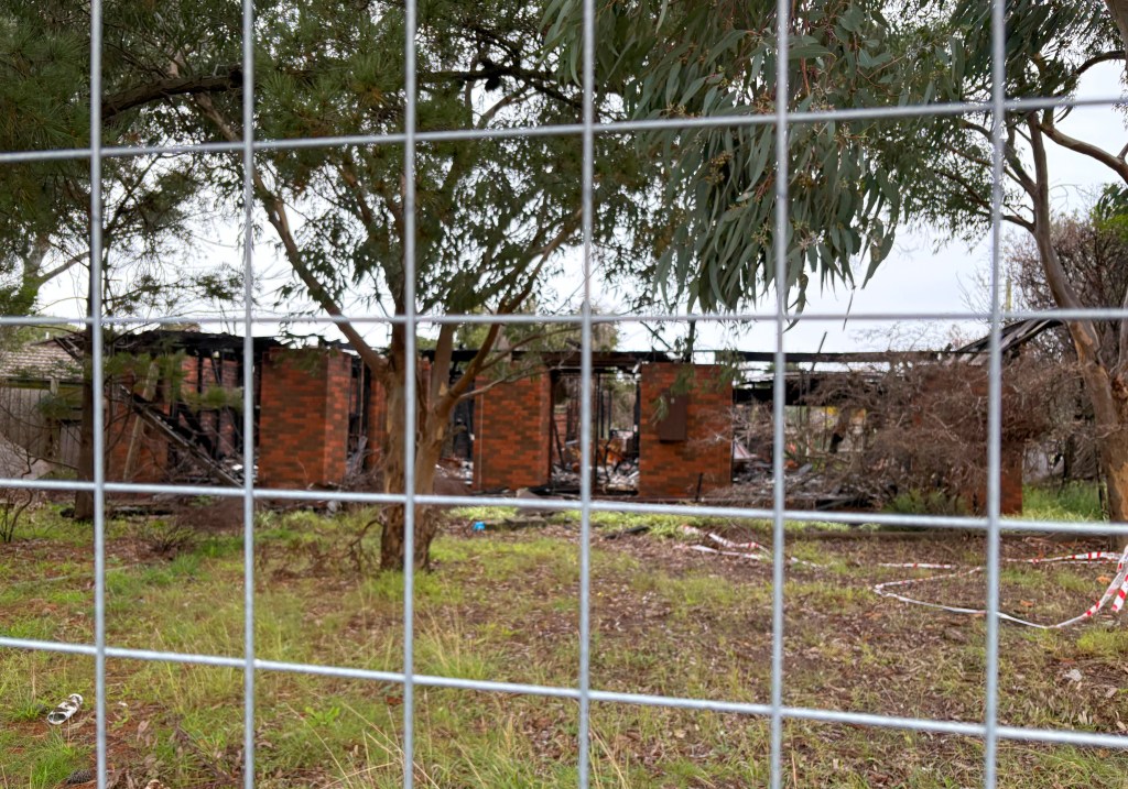 A view of a burnt-out house through a wire fence, surrounded by trees and overgrown grass.