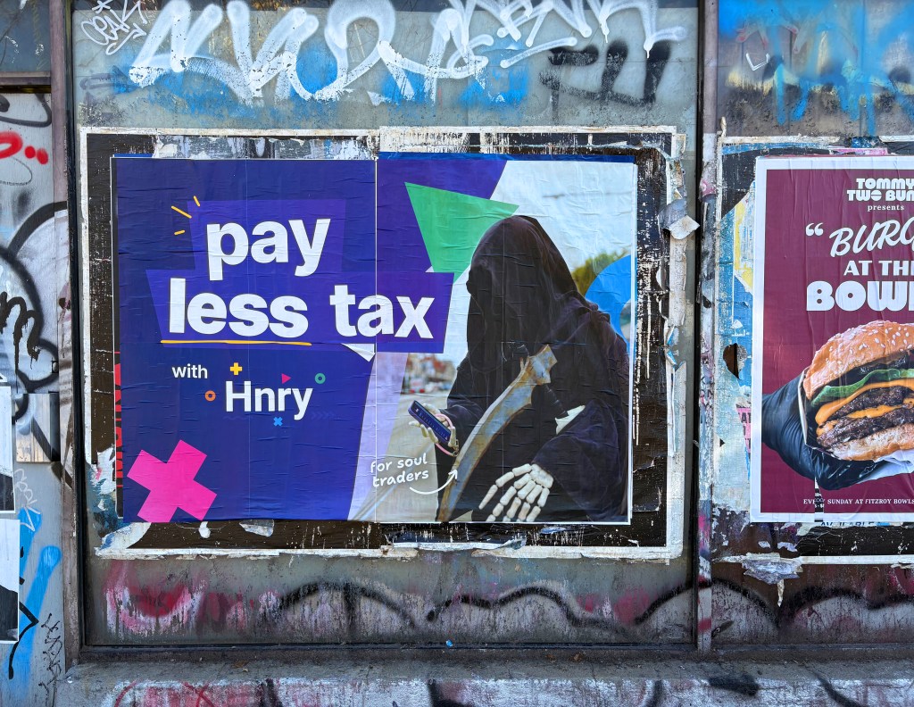 A colourful poster promoting tax savings with 'Hnry' for soul traders, featuring a grim reaper-like figure holding a smartphone, displayed on a graffiti-covered wall.