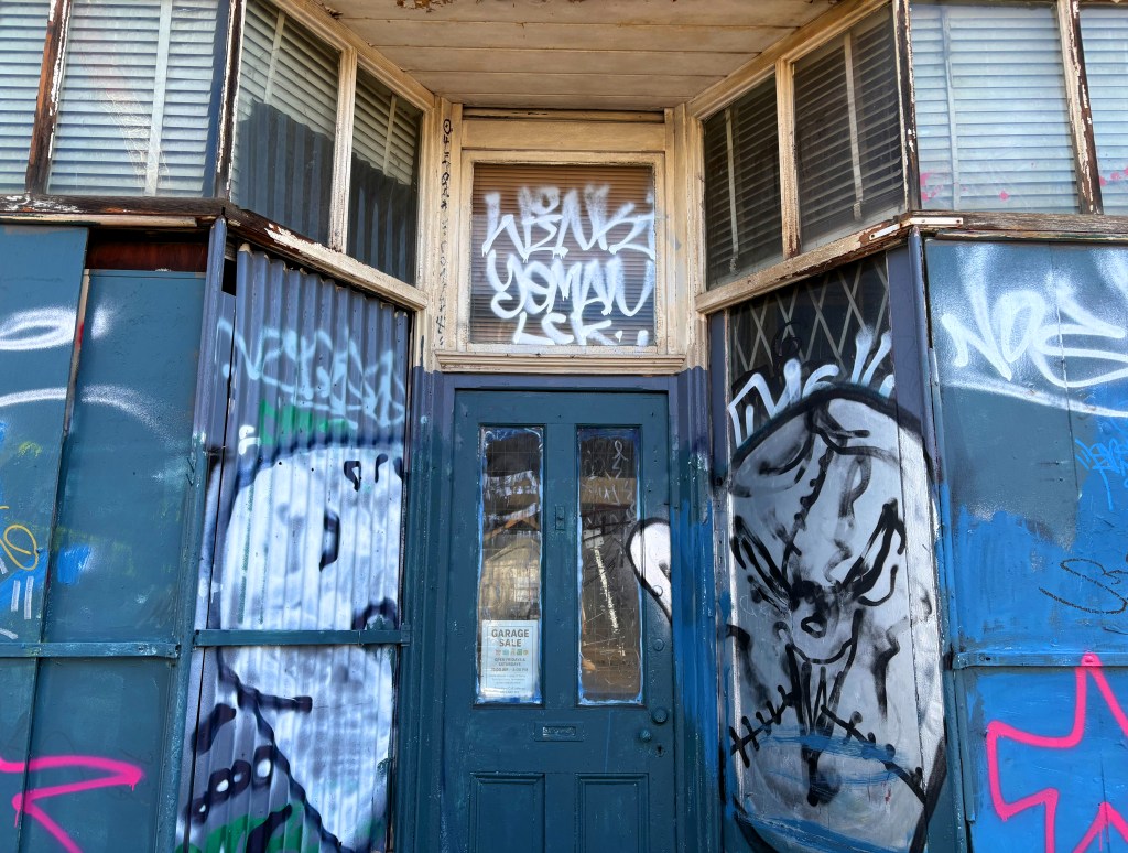 The entrance of a building covered in vibrant graffiti, featuring a mix of artistic tags and illustrations on blue metal and wooden surfaces.