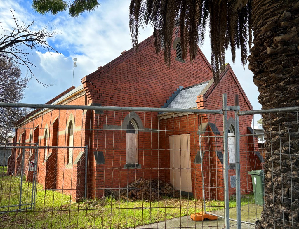A brick building with arched windows, surrounded by a wire fence, set against a cloudy sky. There is a palm tree on the right and a green bin nearby.