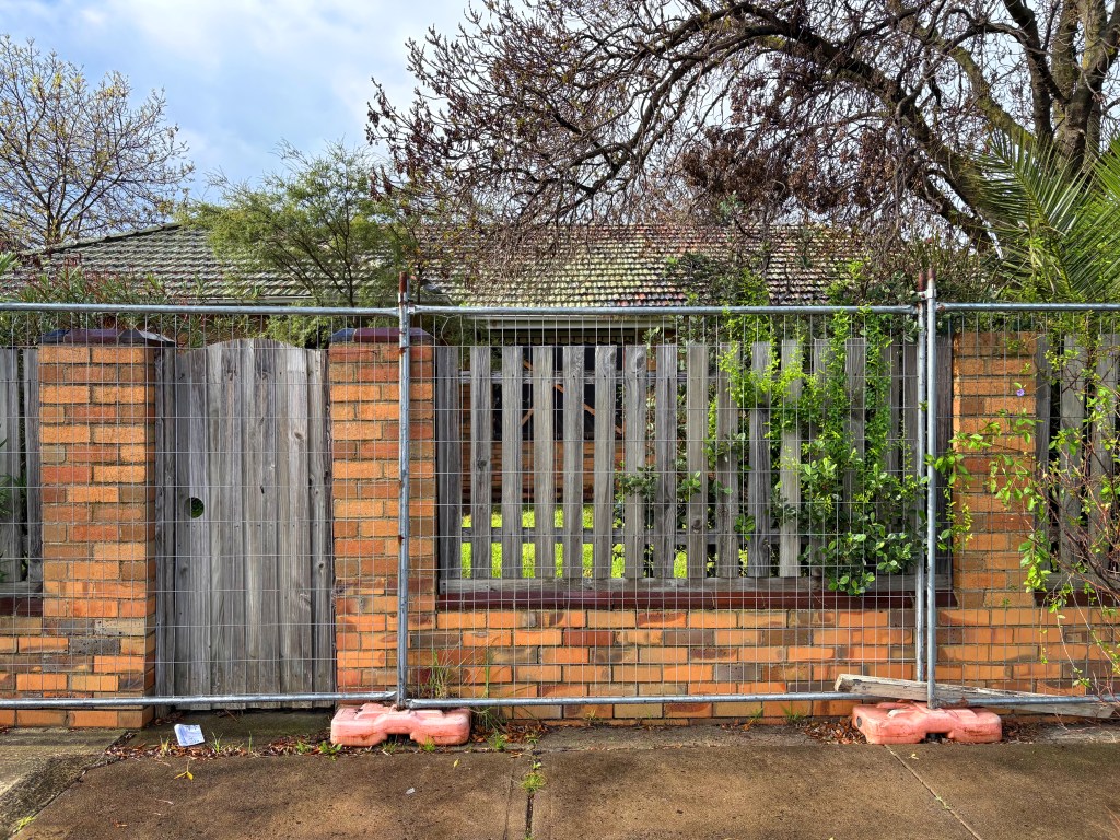 A fenced area with a wooden gate and a brick wall, surrounded by trees and plants, showing a glimpse of greenery behind the fence.