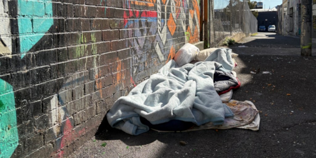 A sleeping area on a pavement, featuring blankets and pillows, set against a graffiti-covered brick wall in an alley.