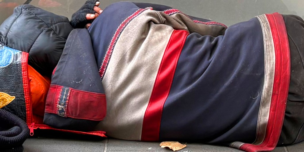 A person lying curled up on a bench, wrapped in colourful blankets and clothing.