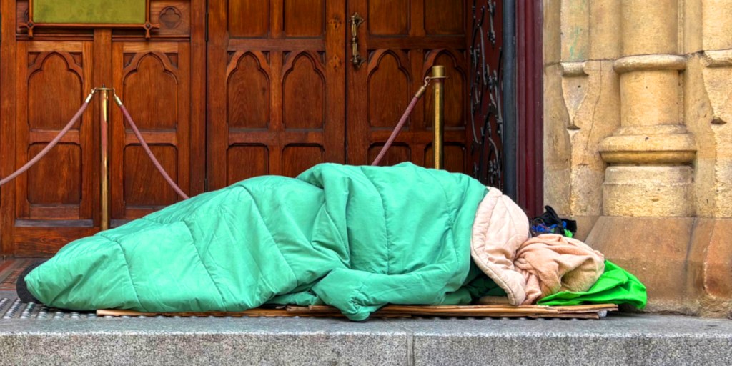 A person sleeping wrapped in a green sleeping bag on a doorstep, with a wooden door and stone wall in the background.