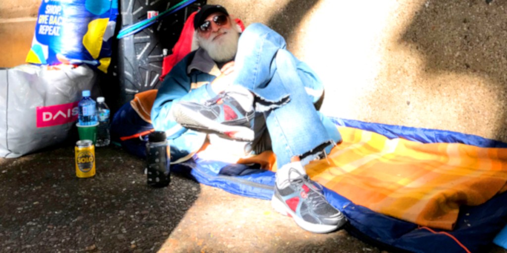 An elderly man with a white beard sitting on a sleeping bag, wearing casual clothes and sunglasses, with water bottles and a drink can nearby.