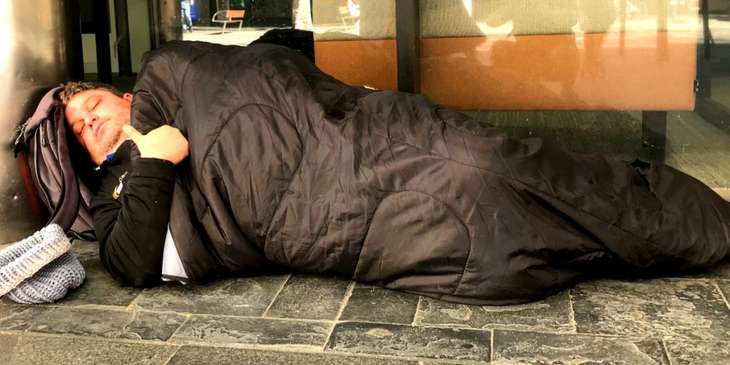 A man sleeping on the pavement, wrapped in a black blanket, with a bag beside him.
