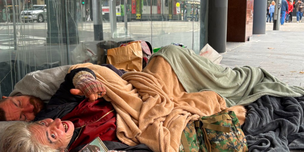 A man and a woman wrapped in blankets, sitting together on the pavement under a glass canopy. The woman is smiling while the man is resting next to her. There are bags and belongings scattered around them.