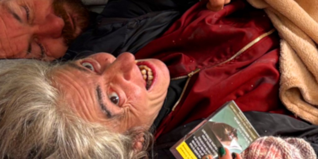 A close-up of two individuals lying together, with one person smiling widely and displaying visible teeth, while the other appears to be asleep. The smiling person has grey hair and is holding a card in their hand.
