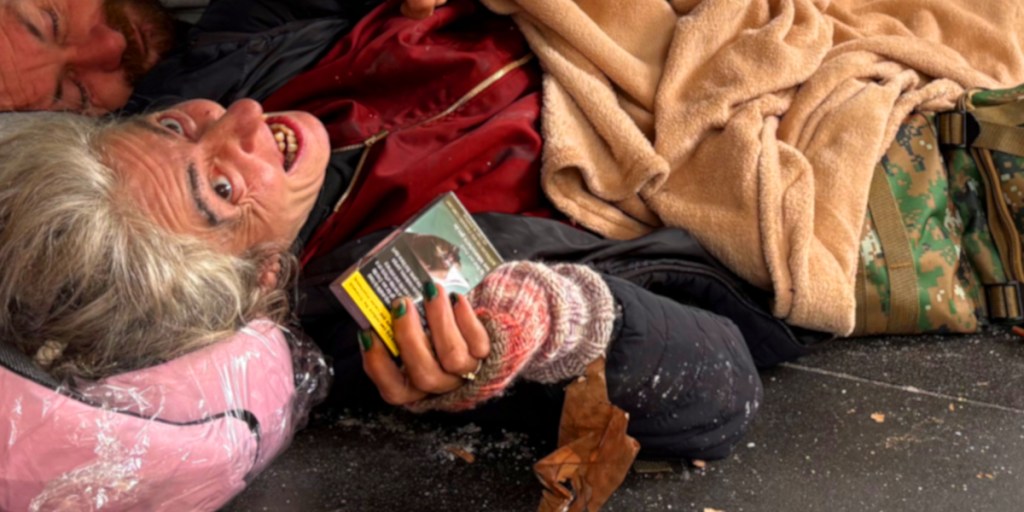 A woman with grey hair and a joyful expression lies on the ground, holding a box. She is wrapped in blankets next to a man, both appearing to be in a homeless setting.