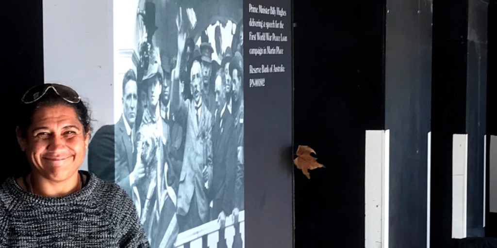 A smiling woman stands in front of a historical black and white mural depicting a crowd, including a man waving and a woman in a hat, with accompanying text about Prime Minister Billy Hughes delivering a speech.