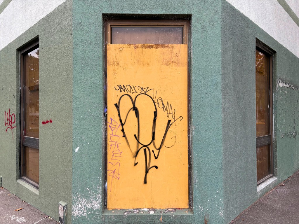 A corner of a building featuring a partially boarded-up window with a yellow door, obscured by graffiti, against a green-painted wall.