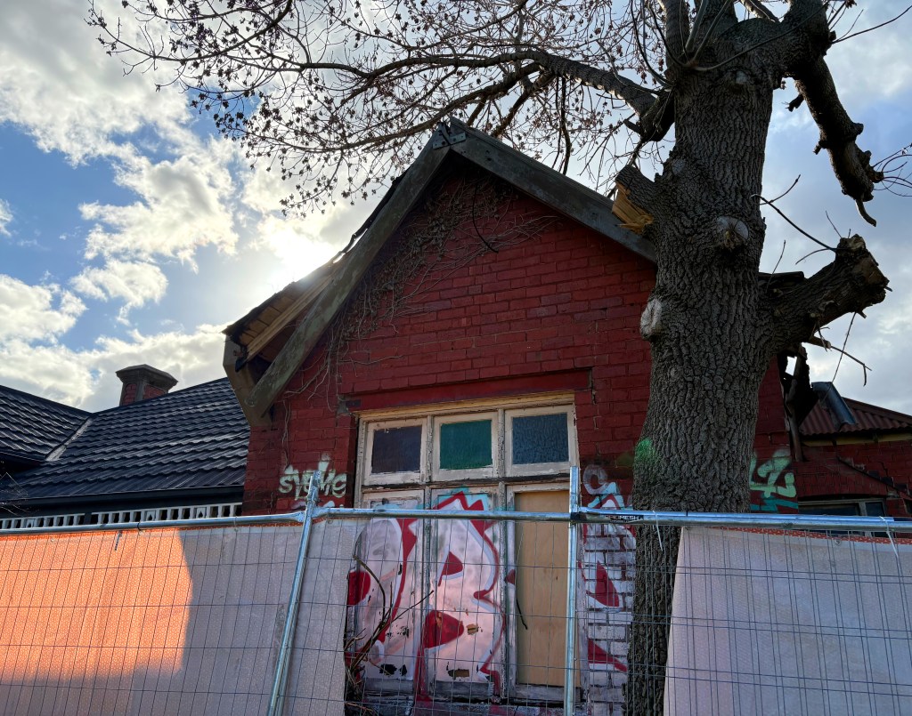 Abandoned red brick house with graffiti, partially covered by a fence and a tree in the foreground, against a cloudy sky.