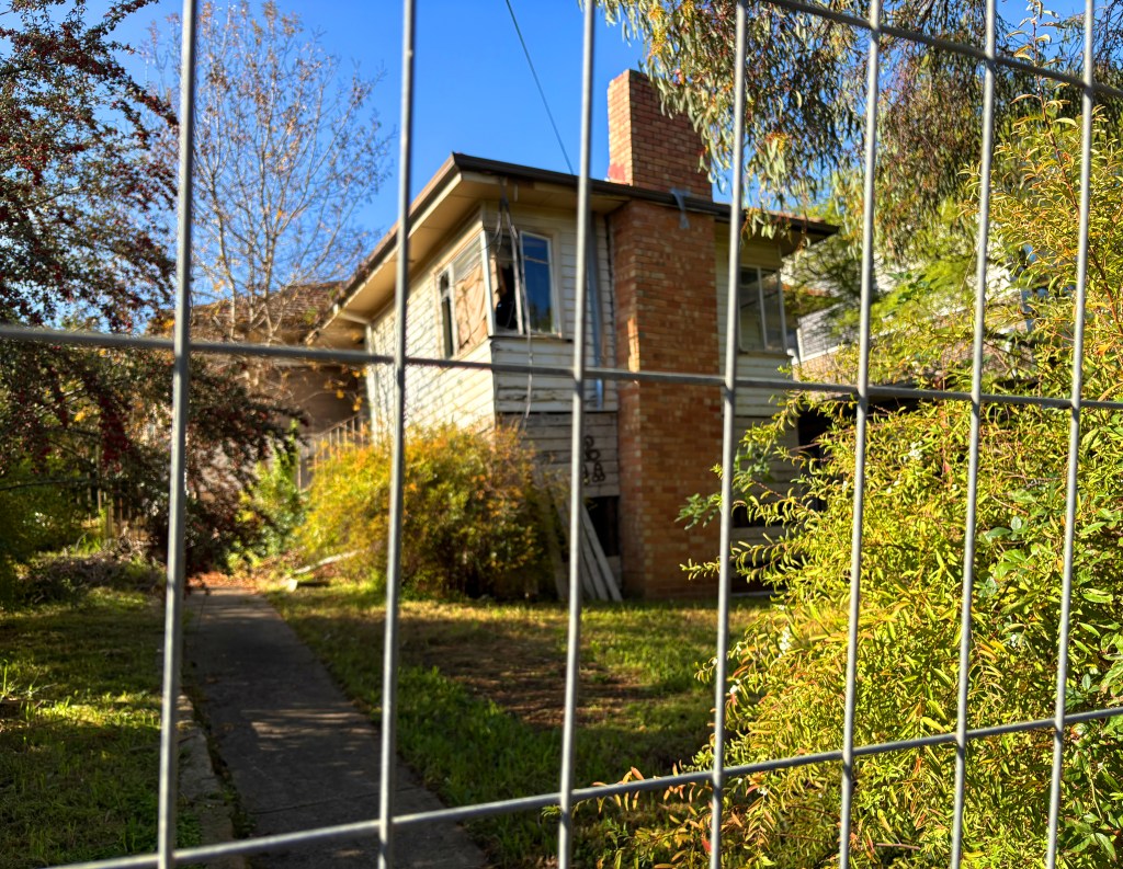 View of a house partially obscured by a fence, surrounded by overgrown vegetation and trees.