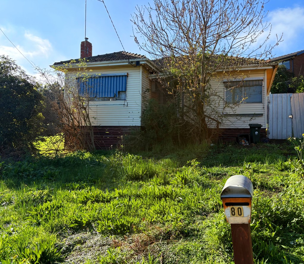 A modest, single-storey house with a partially overgrown garden, featuring a mailbox displaying the number 80 in the foreground.