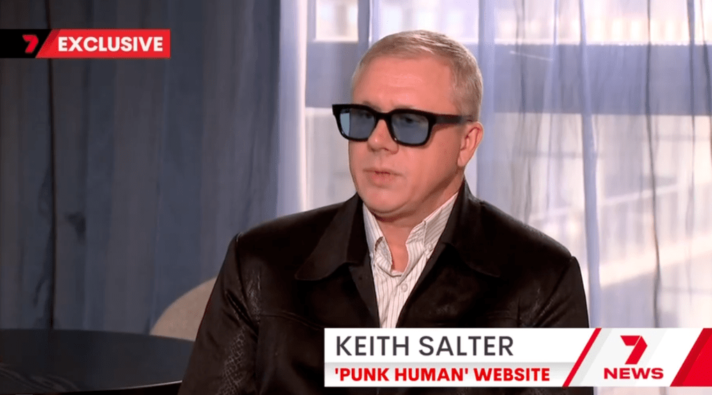A man wearing sunglasses and a black leather jacket is seated, engaged in an interview. The background features soft curtains and a modern interior setting. Text overlay reads 'EXCLUSIVE' and includes the name 'KEITH SALTER' and the title 'PUNK HUMAN' WEBSITE'.