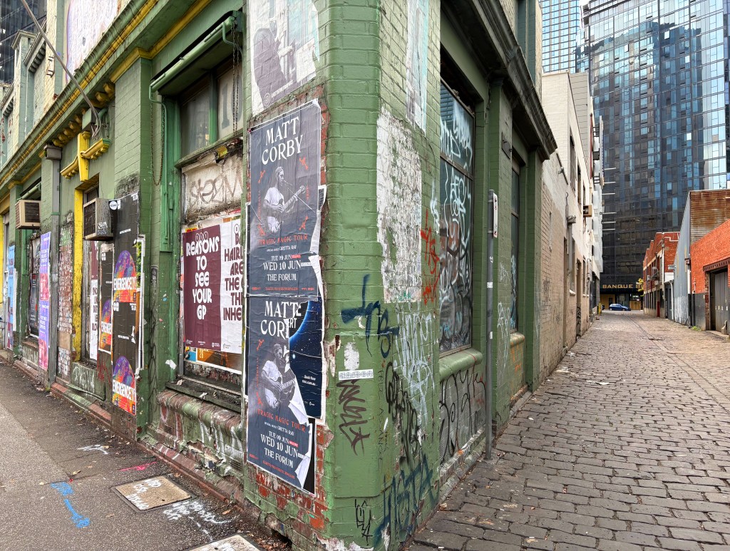 A colourful urban alley featuring an old building covered in posters and graffiti, with cobblestone pavement and modern buildings in the background.