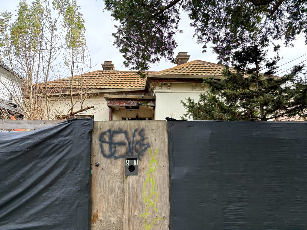 A partially hidden house behind a wooden fence with black tarpaulin and graffiti, surrounded by trees.