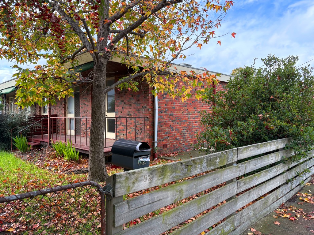 An autumn view of a brick house with a wooden porch, surrounded by trees and shrubs. A black mailbox sits by a wooden fence, with fallen leaves on the ground.