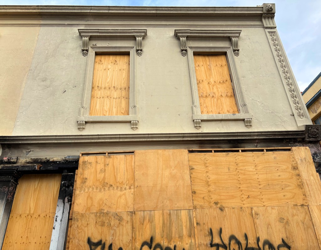 A building facade with two boarded-up windows, featuring ornamental elements above the windows and plywood covering the lower portion, with graffiti visible on the boards.