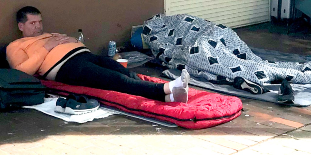 A man lying on a red sleeping mat on the street, wearing an orange top and black leggings, with a concentrated expression. Beside him, another person is covered with a patterned blanket, partially visible.